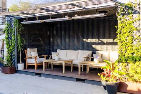 Modern rooftop lounge area with wooden furniture, black shade curtains, and greenery