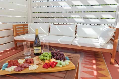 Wooden deck with white cushioned seating, wine bottle, and charcuterie board overlooking green landscape