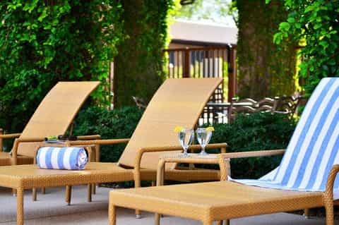 Poolside lounge area with woven chaise lounges, striped towels, lush green hedges, and guest cabanas
