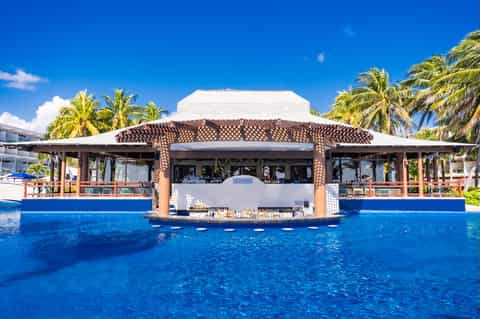 Swim-up bar in tropical resort pool with tiki-style roof, palm trees, and white resort building backdrop