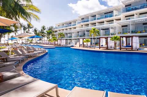 Large resort pool with blue water, white building, cabanas, palm trees, and multiple lounge areas