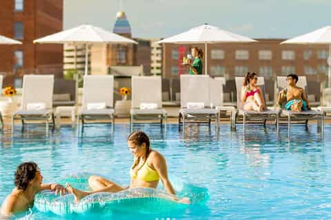Resort guests relaxing in pool with lounge chairs and umbrellas on urban rooftop