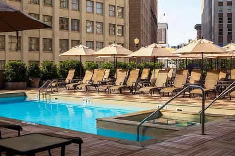 Urban rooftop pool with tan umbrellas, lounge chairs, and city buildings in background