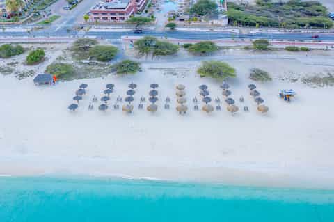 Aerial beach view with rows of dark and tan umbrellas, white sand, turquoise water, and coastal road