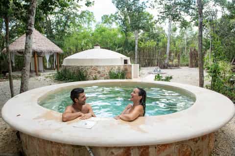 Couple relaxing in circular outdoor hot tub surrounded by forest trees and tiki huts