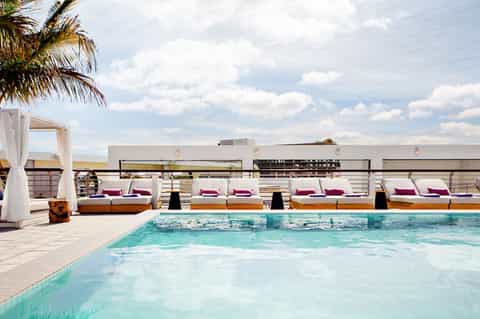 Rooftop pool with turquoise water, lounging areas with pink cushions, white cabanas, and palm trees under blue sky