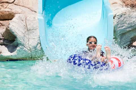 Child splashing down blue water slide into turquoise pool surrounded by rocky cliffs