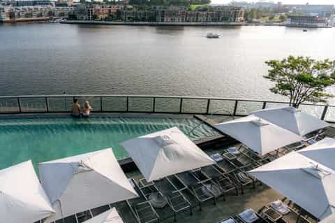 Rooftop infinity pool overlooking river with city buildings, white umbrellas, and waterfront promenade