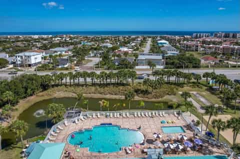 Aerial view of resort pool complex overlooking beach, palm trees, and coastal residential area