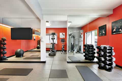 Modern gym interior with red walls, dumbbells, exercise equipment, mirrors, and flat-screen TV