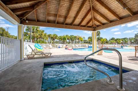 Hot tub under wooden pergola with pool and palm trees visible in background