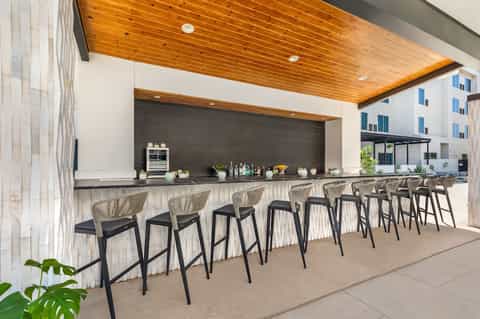 Outdoor bar with wooden ceiling and dark backsplash, featuring woven bar stools and modern design
