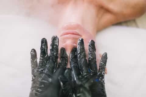 Close-up of hands covered in black spa mud or treatment substance against white towel background.