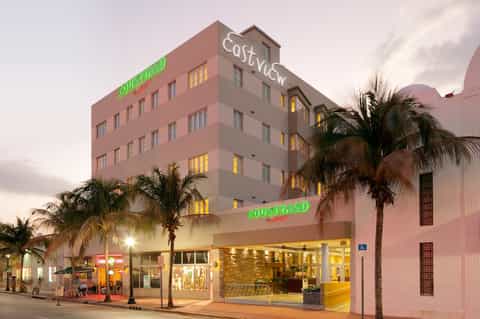 Pink art deco hotel building illuminated at dusk with palm trees and street-level retail
