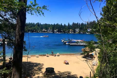 Sandy beach with boats, dock, clear blue water, and tree-lined shoreline with residential homes