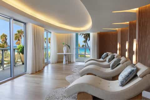 Modern spa lounge with curved loungers, ocean-view windows, palm trees, wooden accents, and ambient lighting