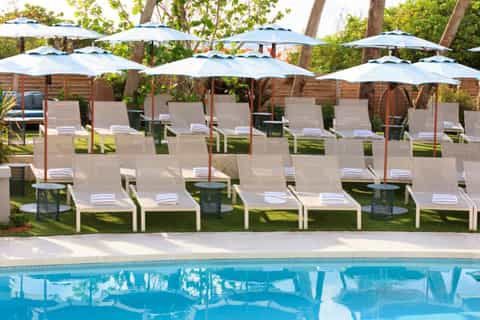 Poolside area with rows of white chaise lounges under blue umbrellas beside sparkling pool