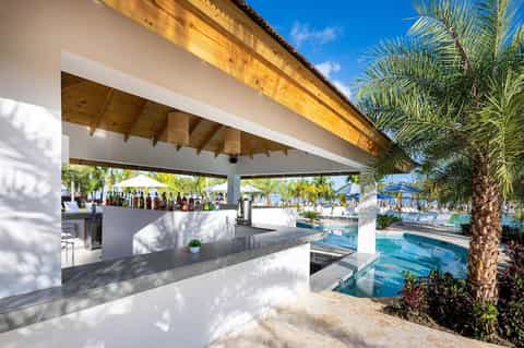 Beachside bar with white counter, wooden pergola, palm trees, and pool view