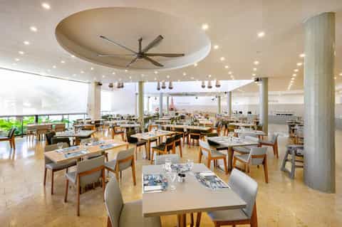 Modern restaurant dining area with wooden tables, white chairs, ceiling fan, and panoramic windows