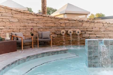 Hot tub with turquoise water, red stone wall backdrop, wooden chairs, and fountain features