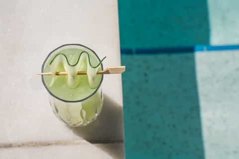 Cucumber water cocktail with wooden paddle garnish on white poolside deck near turquoise water