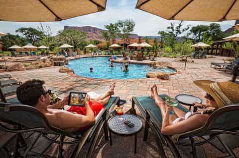 Resort pool area with guests swimming, lounge chairs under tan umbrellas, desert mountains and rock formations visible