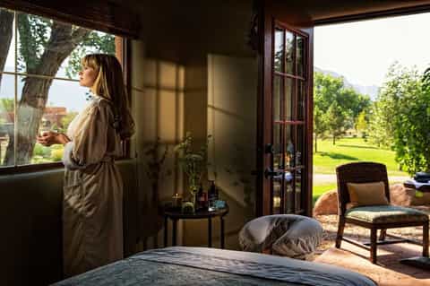 Woman in spa robe looking out at lush green grounds through open glass doors from bedroom