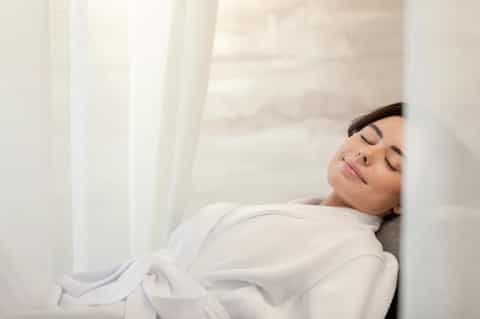 Woman peacefully resting on white bedding in serene, minimalist bedroom setting