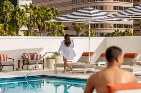 Woman in white shirt walking near modern pool with striped umbrellas and city buildings in background