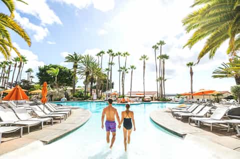 Couple walking into large resort pool surrounded by palm trees, lounge chairs, and orange umbrellas under blue sky