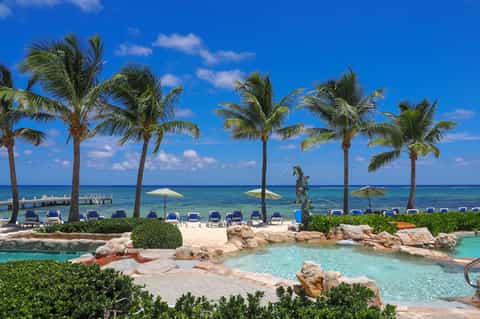 Tropical beachfront pool with palm trees, white umbrellas, turquoise water, blue lounge chairs, and ocean view