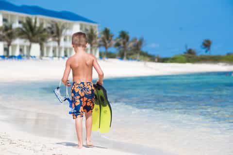 Young boy holding snorkeling fins walking into turquoise tropical ocean water