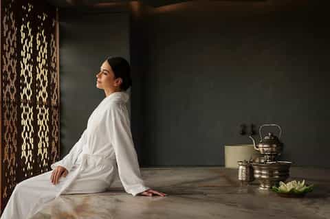 Woman in white robe sitting on marble spa floor with decorative screen and traditional tea service