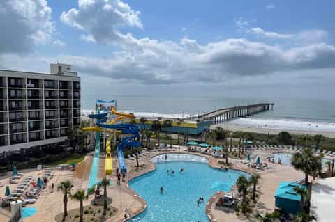 Beachfront resort with large pool, water slide, hotel tower, and ocean pier visible