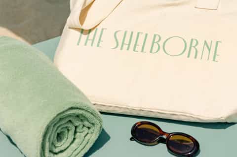 The Shelborne branded canvas tote bag with rolled green towel and sunglasses on turquoise surface