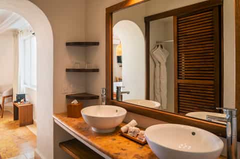Luxury bathroom with dual white vessel sinks on cork countertop, wooden frame mirror, arched alcove, and louvered cabinet