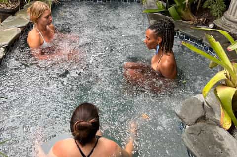 Natural hot spring pool with three women bathing, surrounded by tropical plants and stone edges