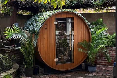 Wooden barrel-shaped sauna pod with curved door, ivy covering, and potted palms in garden setting