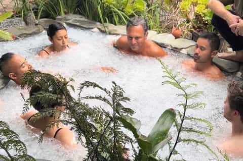 Geothermal hot spring soaking pool with natural vegetation and multiple guests relaxing in steaming mineral water.