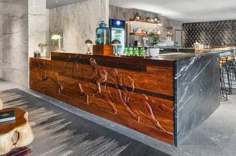 Contemporary hotel bar with carved wooden counter, marble accents, and shelving displays