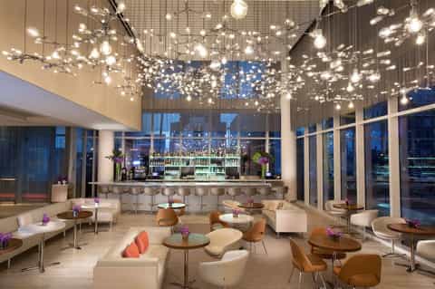 Modern hotel lounge with illuminated bubble ceiling installation, bar area, and seating overlooking city at dusk
