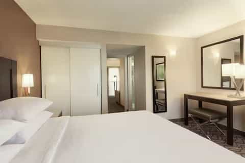 Contemporary hotel room with queen bed, white linens, closet, desk area, and neutral color palette