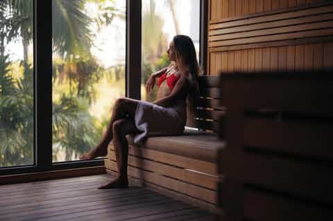 Woman in red swimsuit relaxing in wooden sauna with large windows overlooking tropical garden