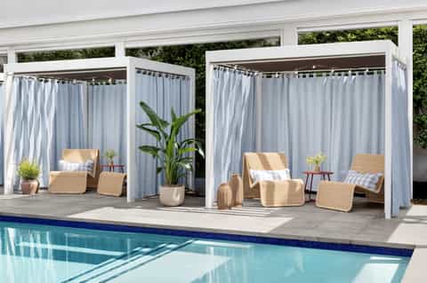 Poolside cabanas with white curtains, woven lounge chairs, potted plants, and blue swimming pool