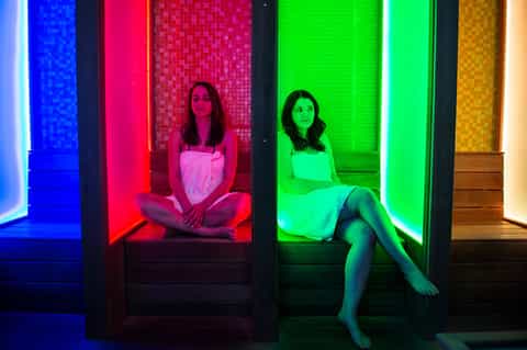 Two women sitting in colorful neon-lit spa pods with blue, red, and green ambient lighting