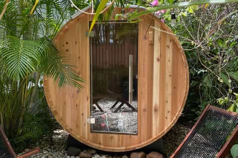 Wooden barrel-shaped sauna cabin nestled in tropical garden with palm fronds and lush vegetation