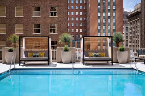 Urban rooftop pool with bright blue water, black cabanas with yellow pillows, and downtown city buildings