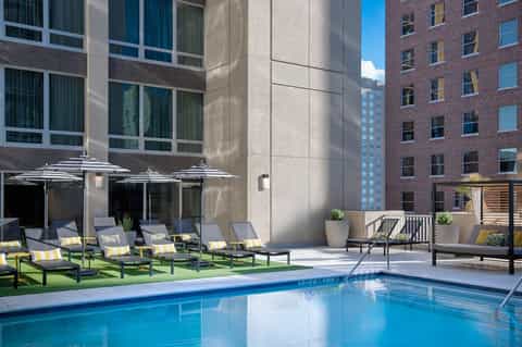 Urban rooftop pool surrounded by striped umbrellas, lounge chairs, and modern city buildings