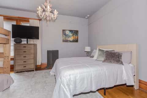 Bedroom with white bed, wooden dresser, TV, pale purple walls, hardwood floors, and crystal chandelier