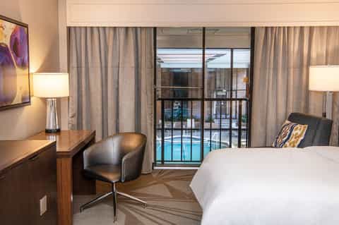 Modern hotel bedroom with bed, desk, chair, and glass doors opening to pool view with floor-to-ceiling curtains